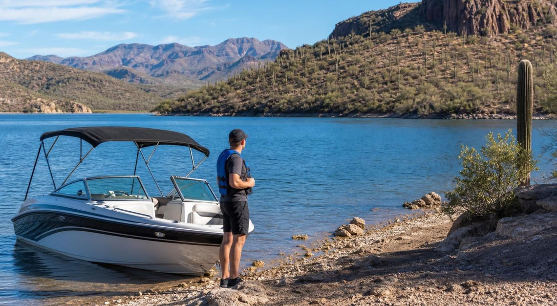 Person wearing a life jacket standing next to a motorboat on a calm Arizona lake with desert mountains in the background.